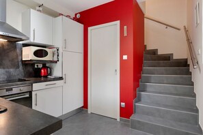 A kitchen with white cabinets, a red wall, a microwave, a kettle, and a staircase.
