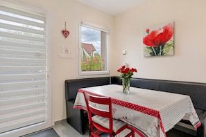 Un coin repas avec une table dressée pour deux personnes, une chaise rouge, un banc, une fenêtre donnant sur la verdure et un interrupteur fixé au mur.