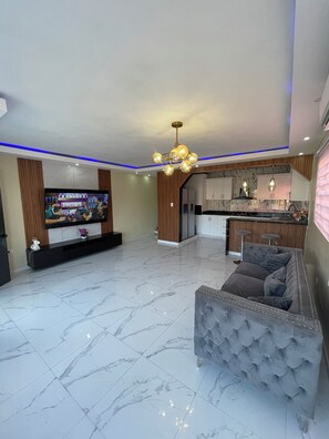 A modern living room with a flat-screen TV, a grey sofa, and a kitchen area with a marble backsplash. - Comfort One-Bedroom Apartment (Santo Domingo)