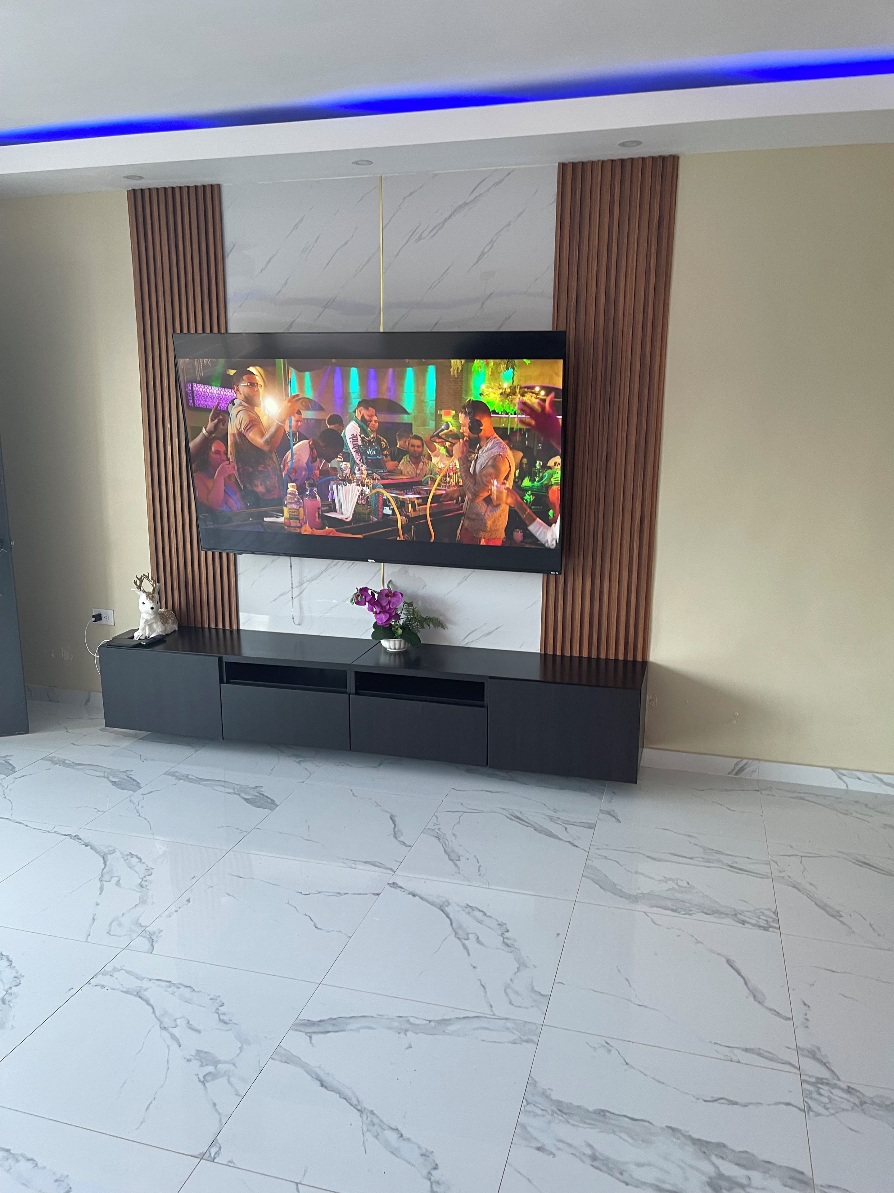 A flat-screen TV mounted on a wall with a wooden panel design, a marble-tiled floor, and a small decorative plant on the entertainment unit.