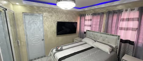 Quarto com cama, mesas de cabeceira, televisão e luminária no teto.