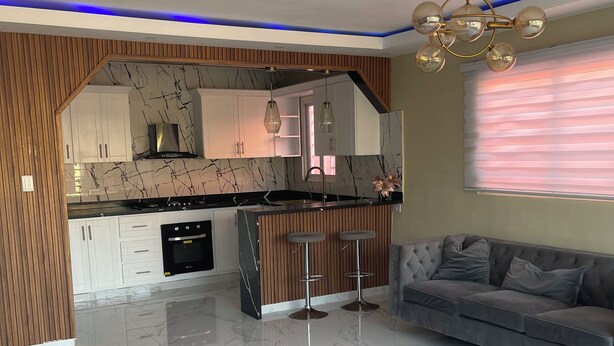 Una cocina moderna con encimeras de mármol, horno empotrado y comedor con sofá gris y dos taburetes de bar.
