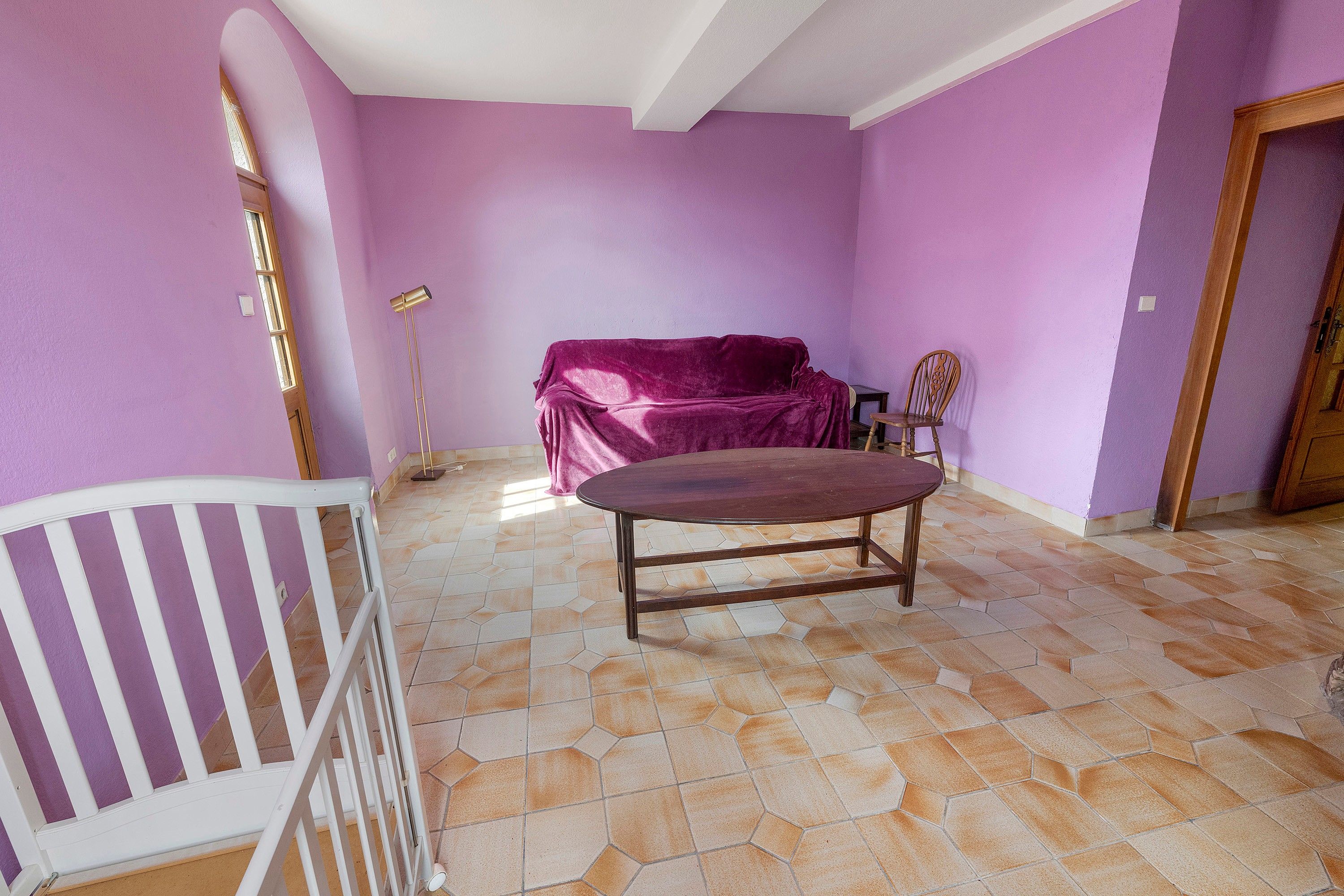Une pièce avec un mur violet, une table ronde, un canapé recouvert d’un tissu, une chaise en bois et un berceau blanc.