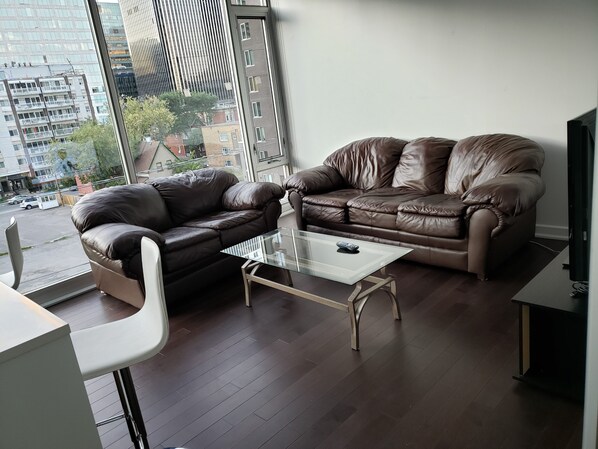 A modern living room with two leather sofas, a glass coffee table, and a view of a cityscape through large windows.