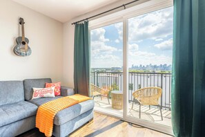 Ein Wohnzimmer mit einem grauen Sofa, einer Gitarre und einem Balkon mit Korbstühlen und einem Tisch.