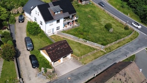 A two-story house with a balcony, a driveway with parked cars, and a road in the background.
