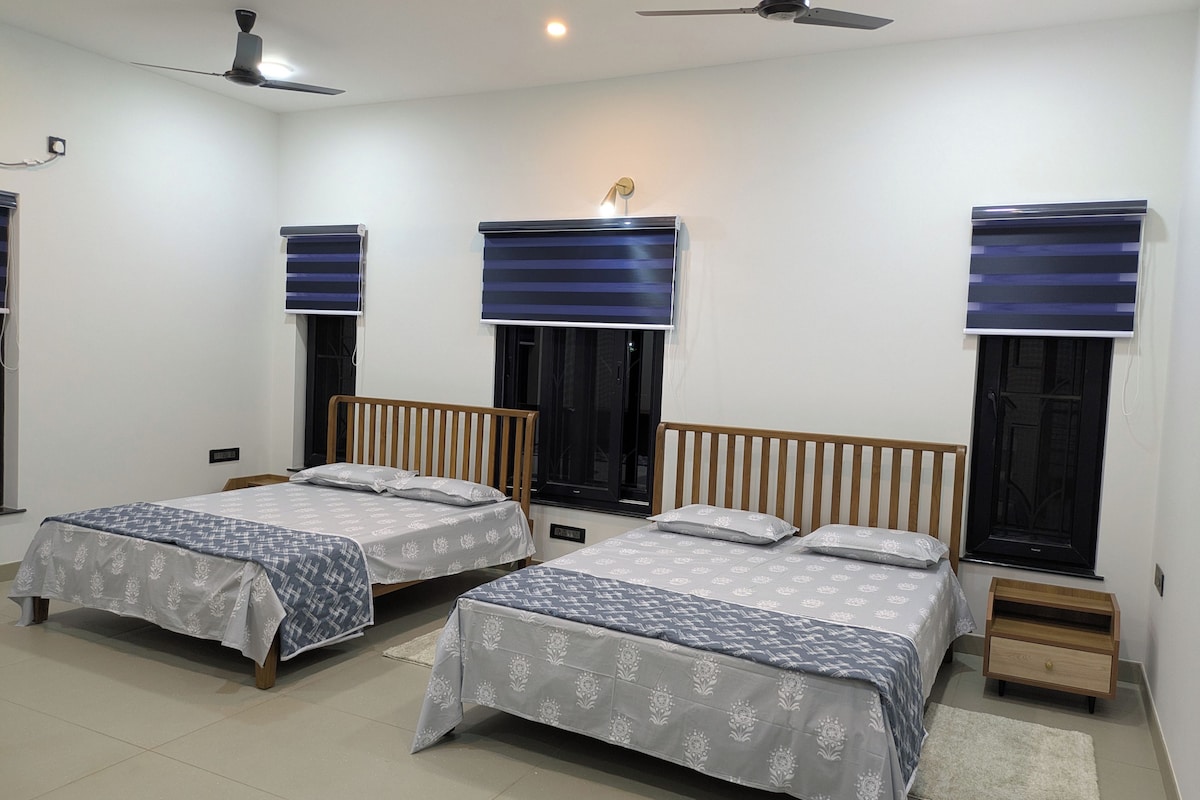 Habitación con dos camas, cada una con cabecera de madera y colcha estampada. Hay ventiladores de techo, una mesita de noche de madera con un cajón y una alfombra blanca en el suelo.