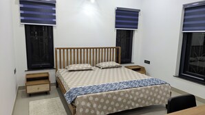 A bedroom with a single bed, bedside tables, a desk, and two windows with blinds. - Abhimaan (Mysore)