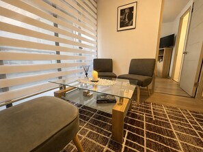 A modern living room with a glass coffee table, two grey armchairs, a brown sofa, a patterned rug, a framed picture on the wall, and a television in the background.