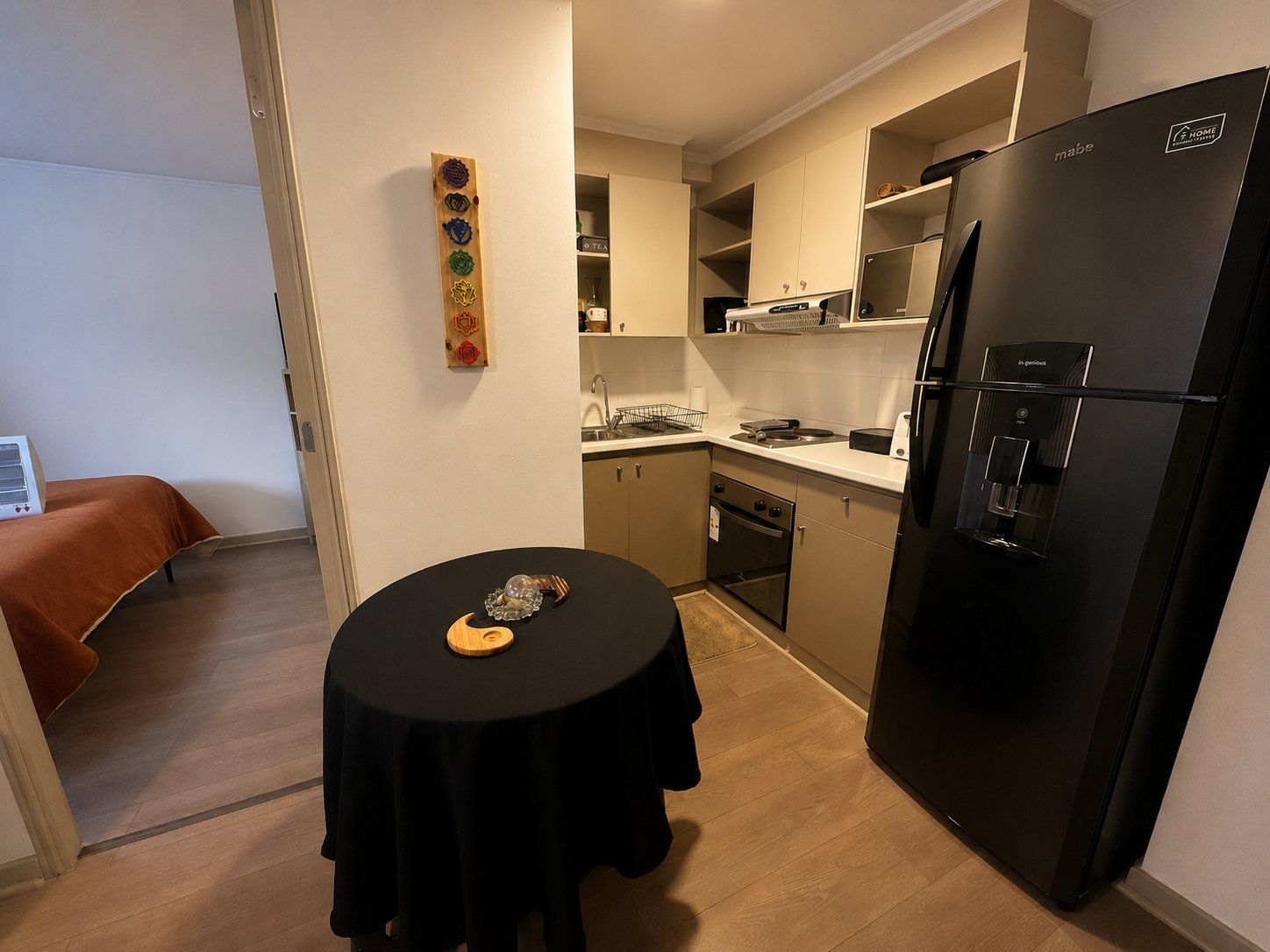 A compact kitchen with a black refrigerator, a small dining table with a placemat, and a wooden decorative piece on the wall.