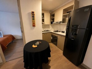 A compact kitchen with a black refrigerator, a small dining table with a placemat, and a wooden decorative piece on the wall.
