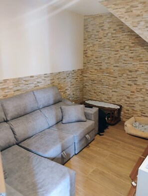 A living room with a grey sectional sofa, a small table with a lace tablecloth, a pet bed, and a stone-patterned wall.