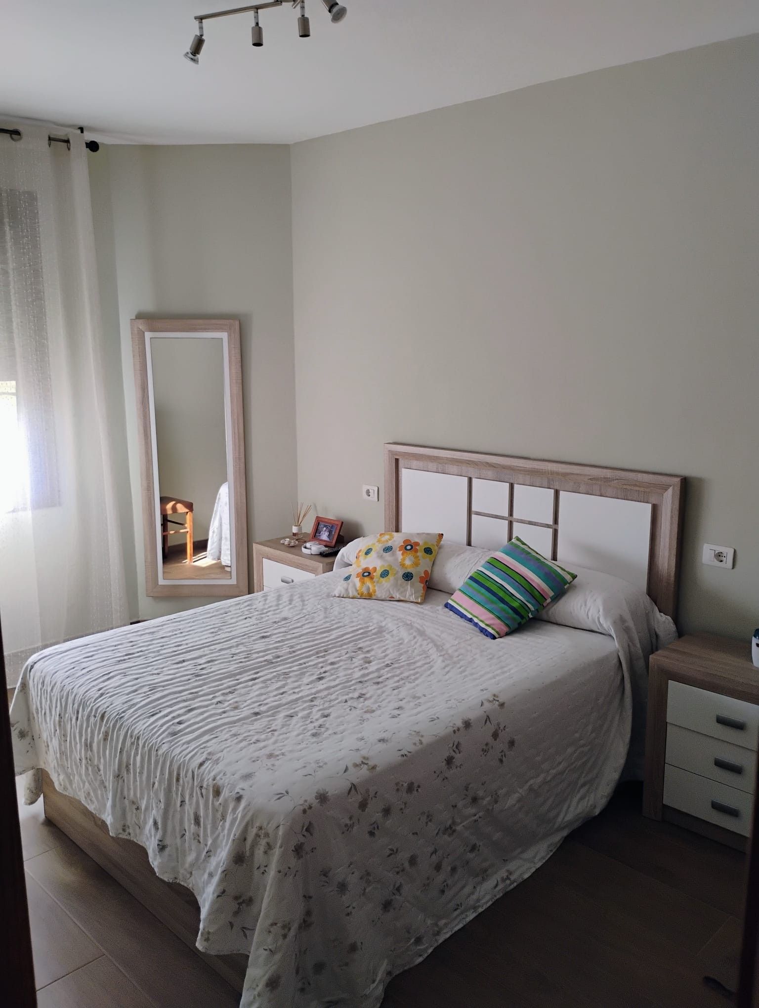 Un dormitorio con cama, mesita de noche, espejo y ventana con cortinas.
