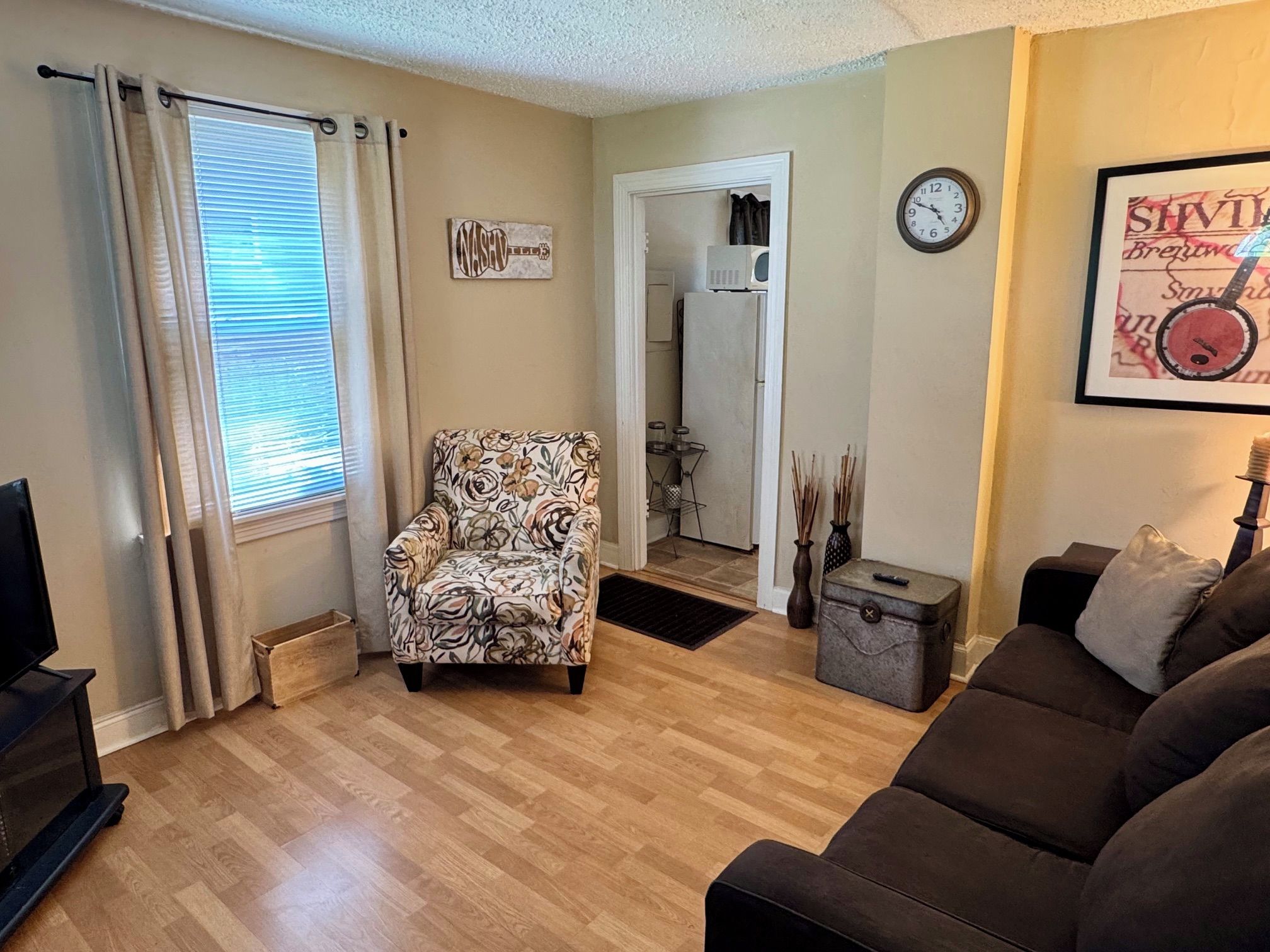 A living room with a floral armchair, a brown sofa, a small table, a clock, and a framed picture on the wall.