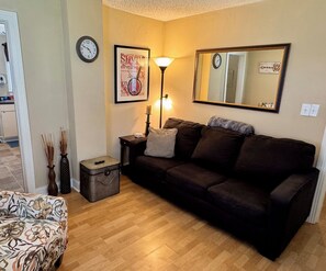 Ein Wohnzimmer mit einem dunkelbraunen Sofa, einer Lampe, einem gerahmten Bild, einer Uhr und einem kleinen Tisch mit einer Vase und einer Schachtel.