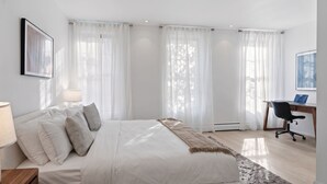 A modern bedroom with a large bed, a desk with a laptop, and a chair. - Stylish 850 SF SoHo 1BR W/D (New York)