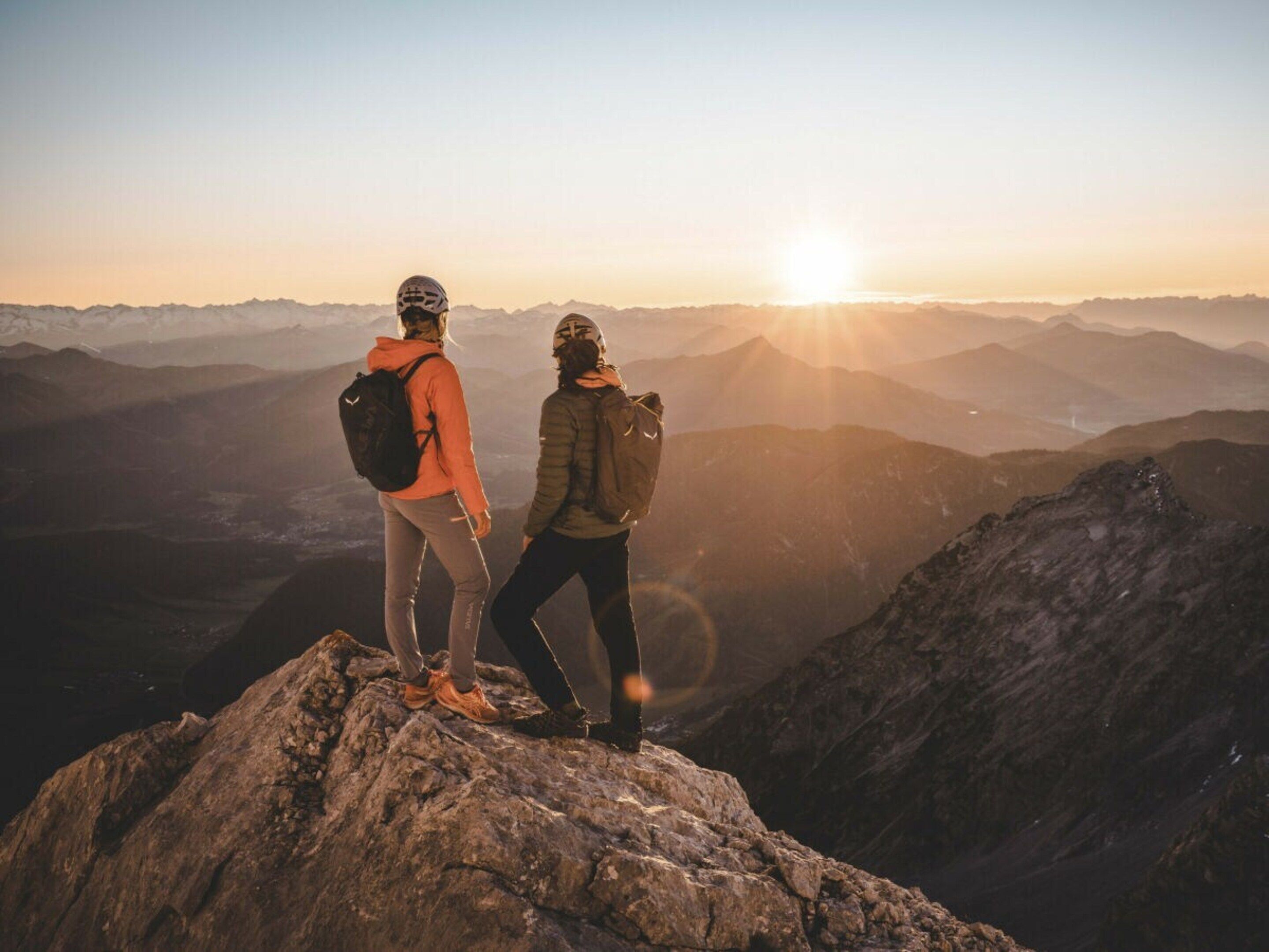 Two people standing on a rocky peak overlooking a mountain range with the sun setting in the background.