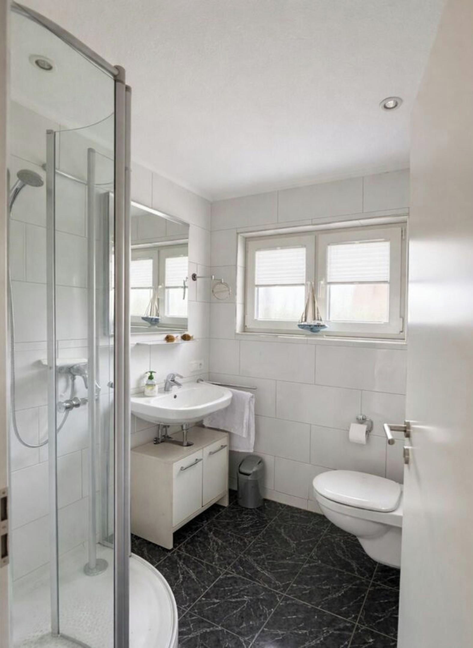 Ein modernes Badezimmer mit Duschkabine aus Glas, einem weißen Waschbecken, einer Toilette und einem Fenster.