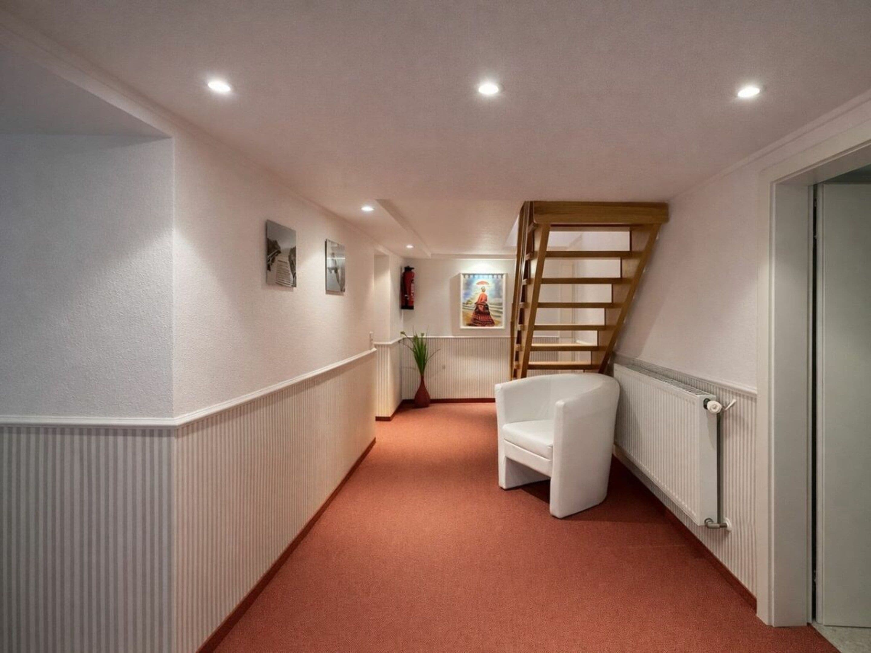 Un pasillo estrecho con un radiador blanco, una silla blanca y una escalera de madera.
