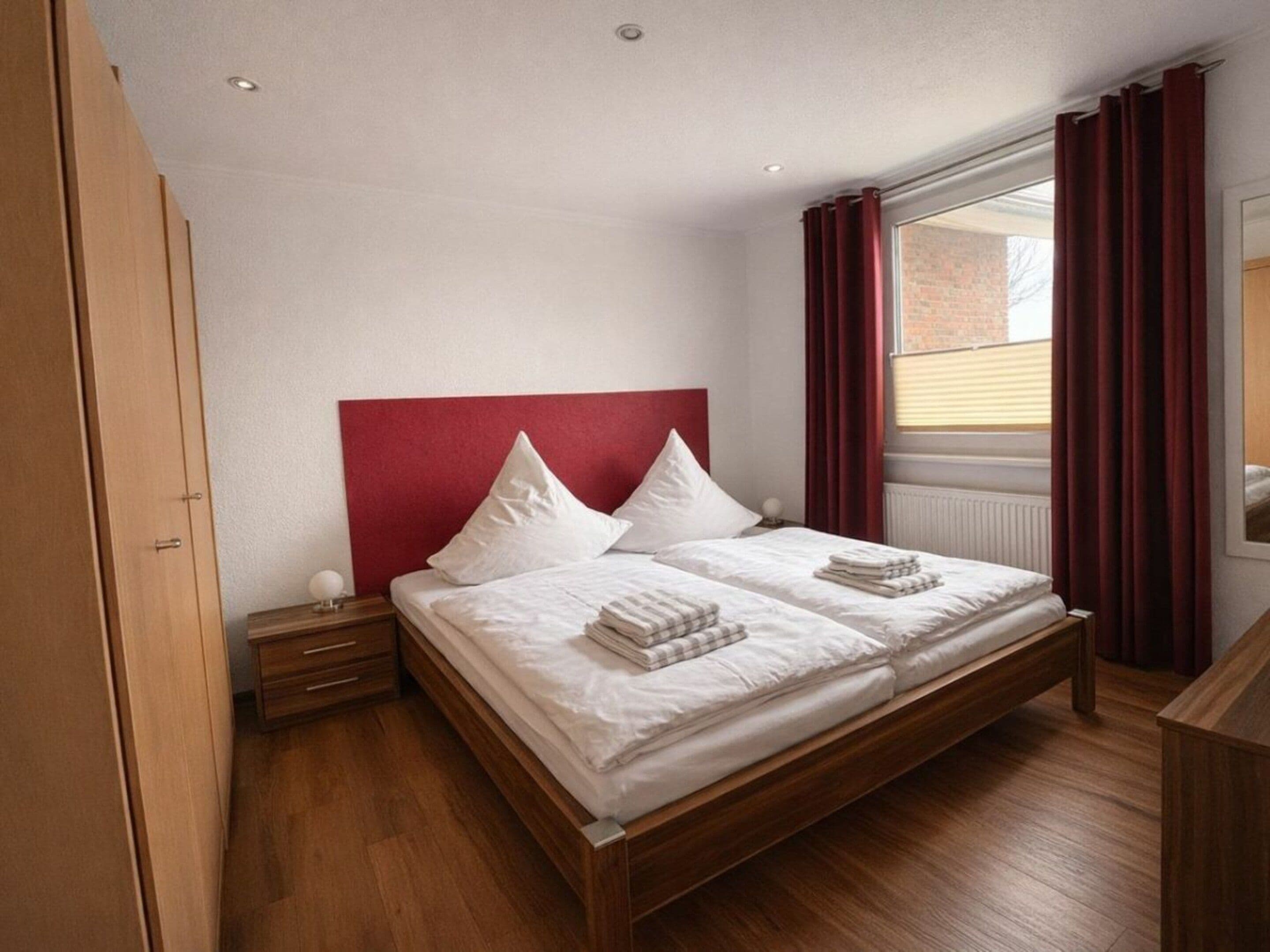 Una cama doble con cabecera roja, dos mesitas de noche, un armario de madera y una ventana con cortinas.