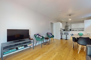A modern living room with a flat-screen TV, green chairs, a wooden dining table, and a kitchen in the background. - Bright 3BR Flat w/Private Garden-Near Station (London)