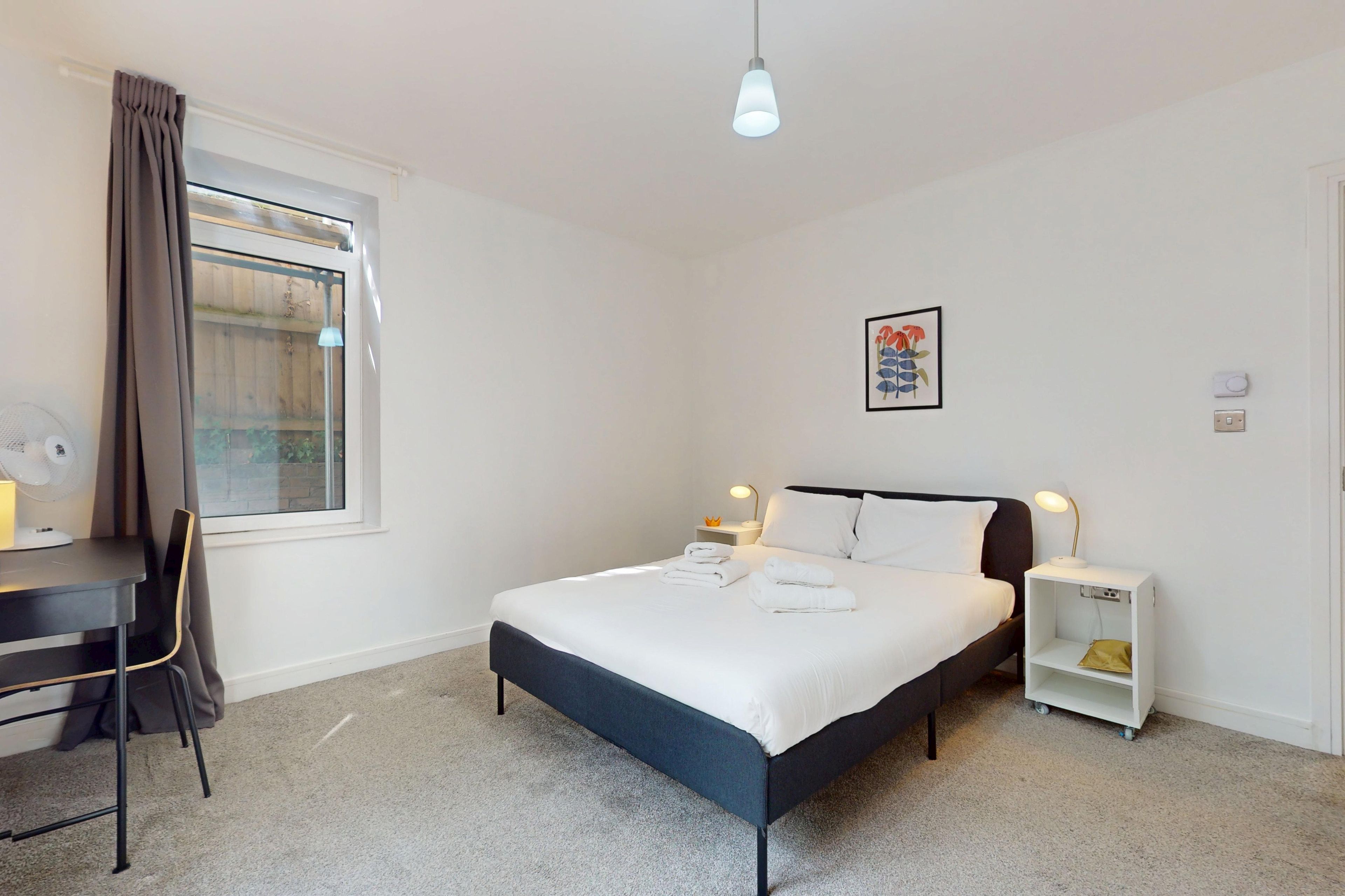 A bedroom with a bed, bedside tables, a desk, a chair, a window with curtains, and a ceiling light fixture.