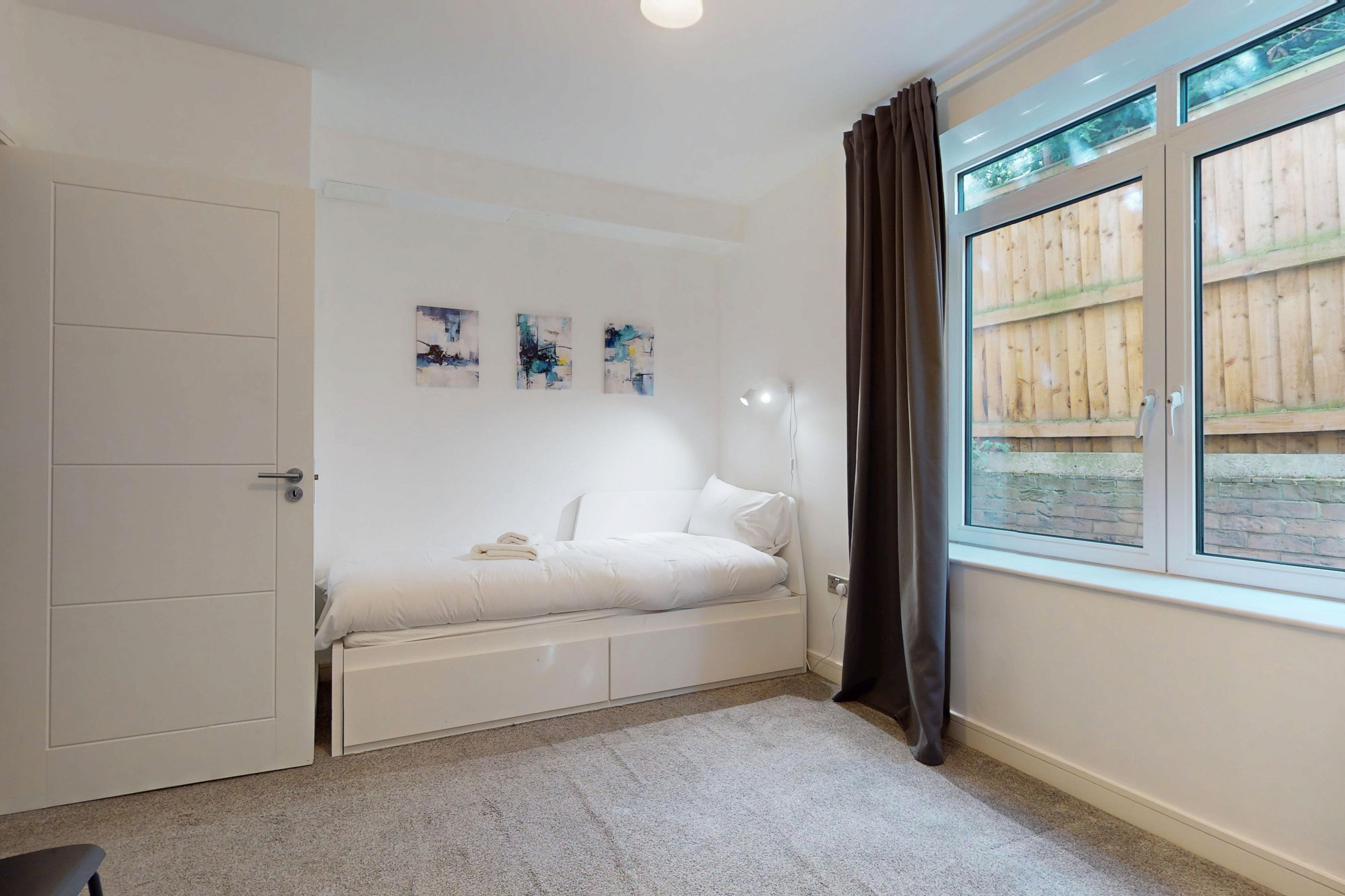 Une chambre à coucher comprenant un lit, une armoire, une fenêtre donnant sur une clôture en bois et une œuvre d’art abstrait accrochée au mur.