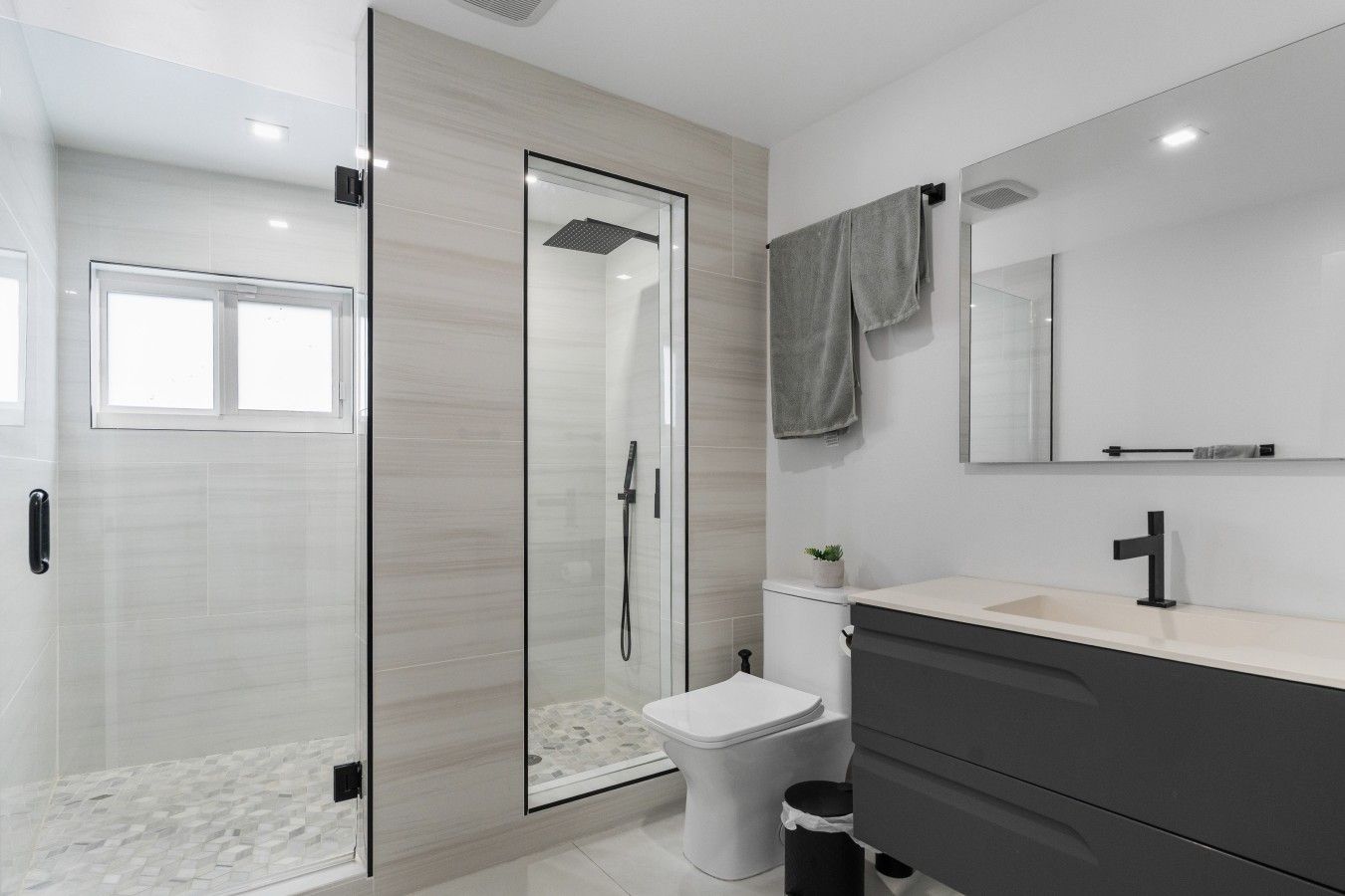 Un baño moderno con ducha envuelta en vidrio, inodoro blanco, tocador oscuro y espejo sobre el lavamanos.