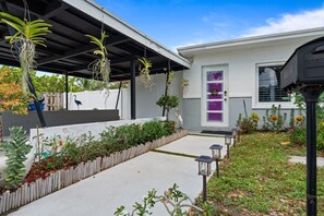 Una casa moderna con un porche cubierto, un camino de concreto y una variedad de plantas y flores.