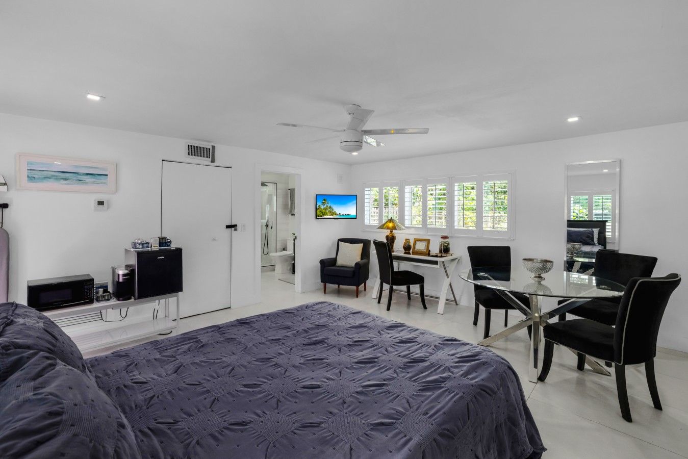 A modern bedroom with a bed, a desk, and a chair. There is a bathroom visible through an open door.