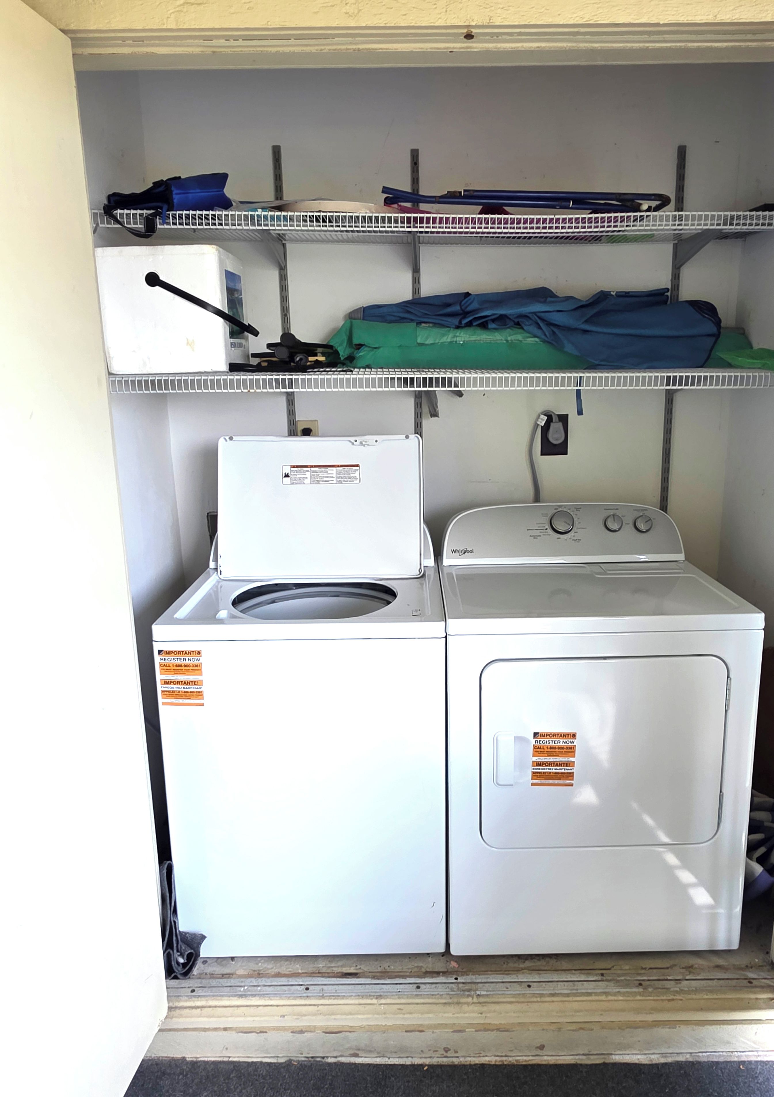 A laundry room with a front-loading washer and dryer, shelving above with folded clothes and cleaning supplies.
