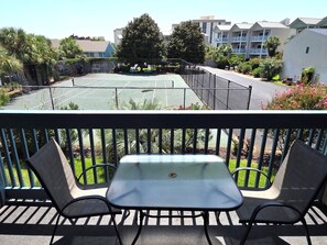 Een balkon met een tafel en twee stoelen, met uitzicht op een tennisbaan en woongebouwen.