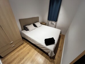Un petit lit bien fait, avec un matelas blanc et deux oreillers, une armoire en bois et une table de chevet avec une lampe.