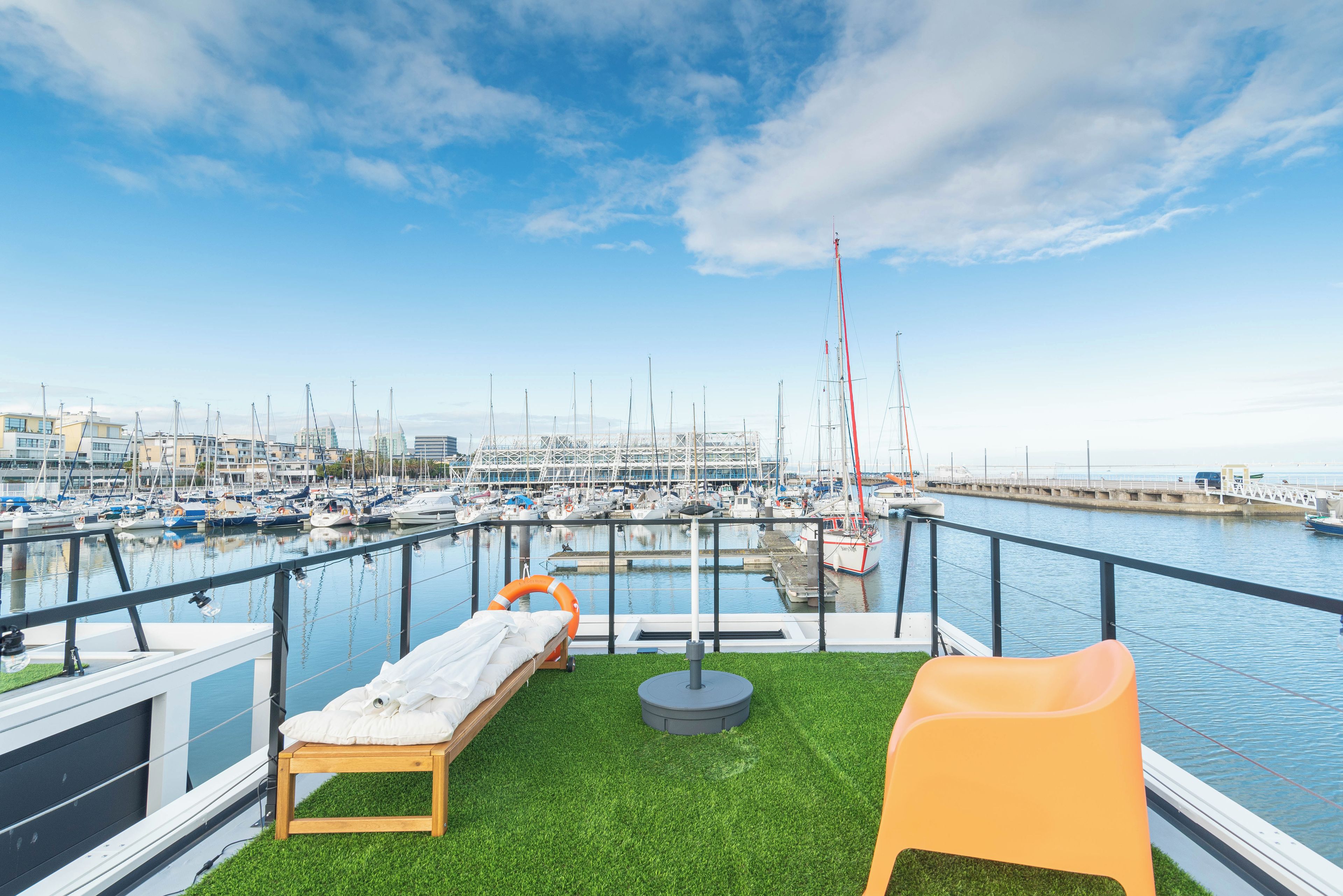 Ein Yachthafen mit vor Anker liegenden Booten, ein Loungebereich mit einer Bank und einem kleinen Tisch sowie ein Blick auf den Hafen und die Gebäude im Hintergrund.