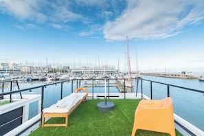 A marina with boats docked, a lounge area with a bench and a small table, and a view of the marina and buildings in the background.