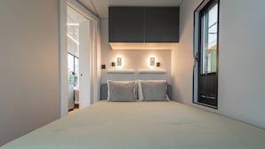 Quarto com cama, duas mesas de cabeceira, janela com persianas e porta que dá para outro cômodo.
