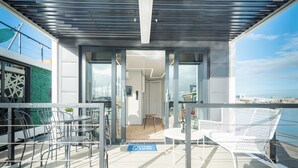 Uma área de estar moderna ao ar livre, com cadeiras e mesas de metal, porta de entrada de vidro e vista para o mar.