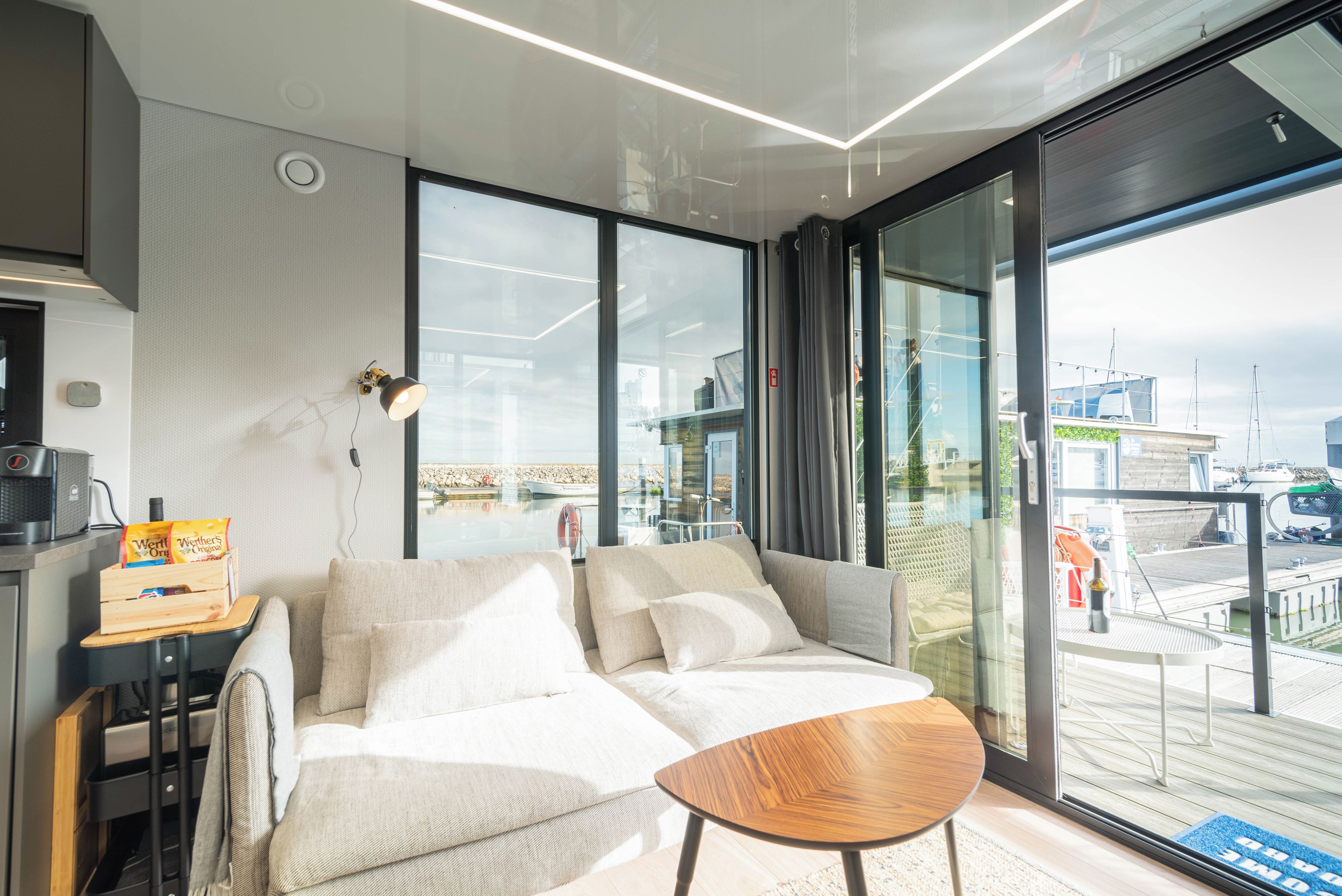 A modern living room with a sofa, a small wooden table, and a large window offering a view of the sea.