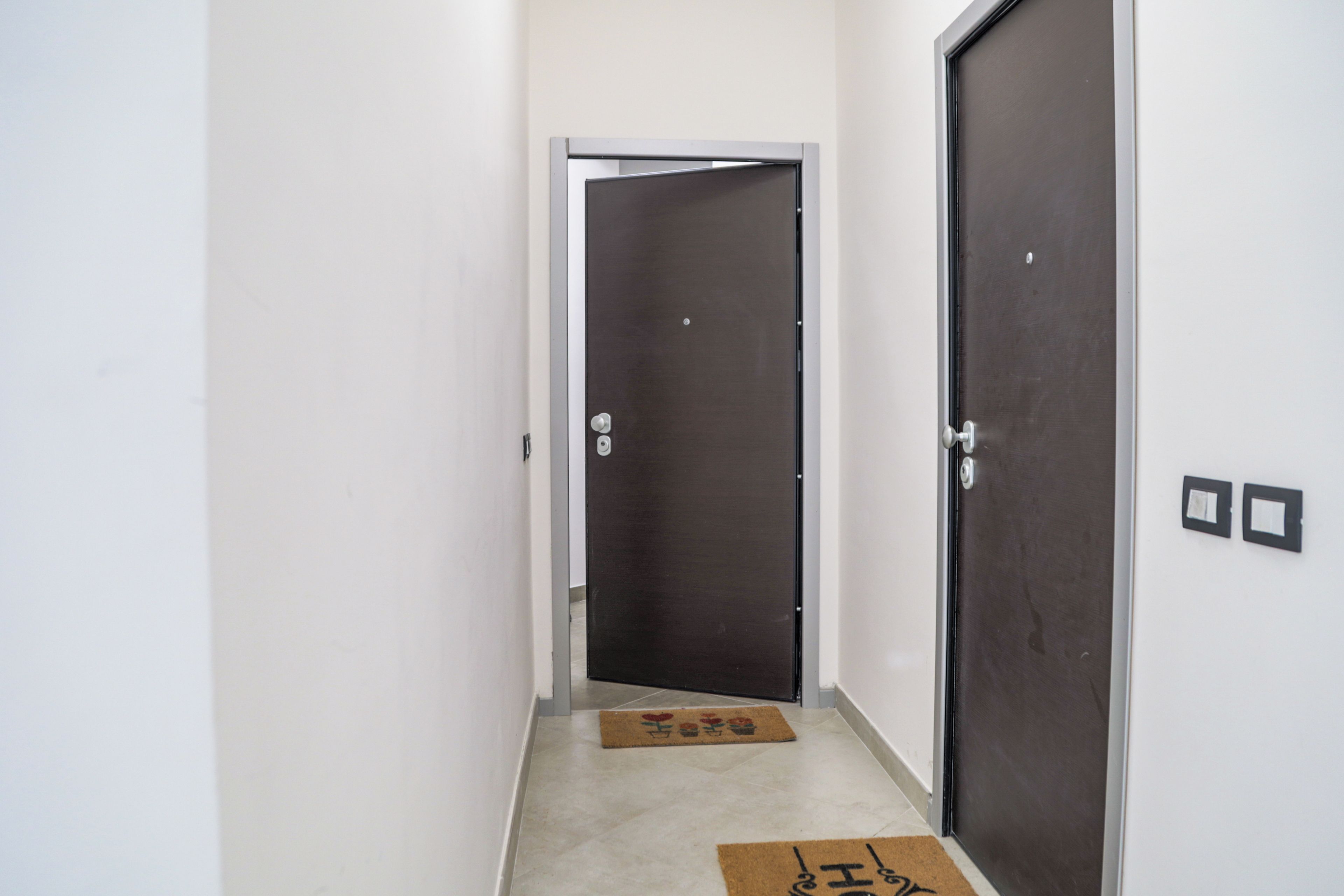 A narrow hallway with two doors, one closed and one open, leading to a room. There are two doormats on the floor.