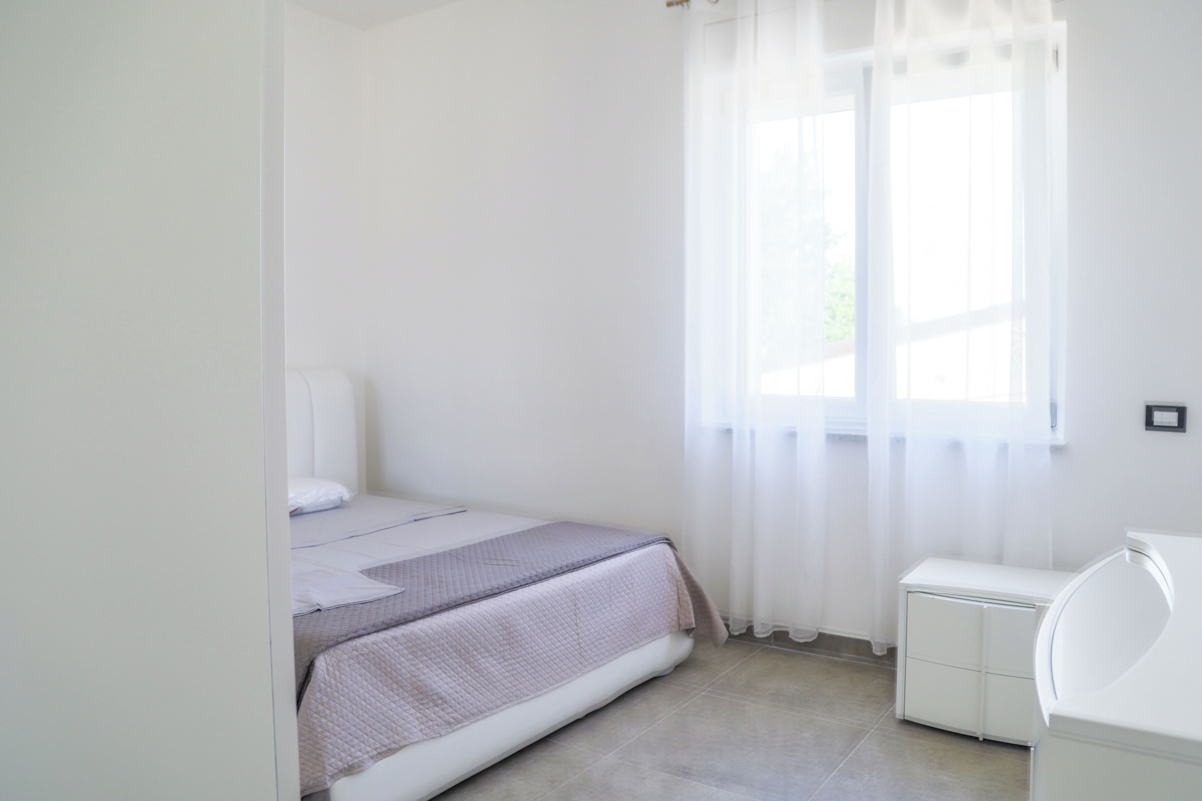 A small, clean bedroom with a bed, a window with sheer curtains, and a white bedside unit.