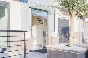 Entrada de un edificio moderno con puerta de cristal, reja metálica y un árbol en maceta.