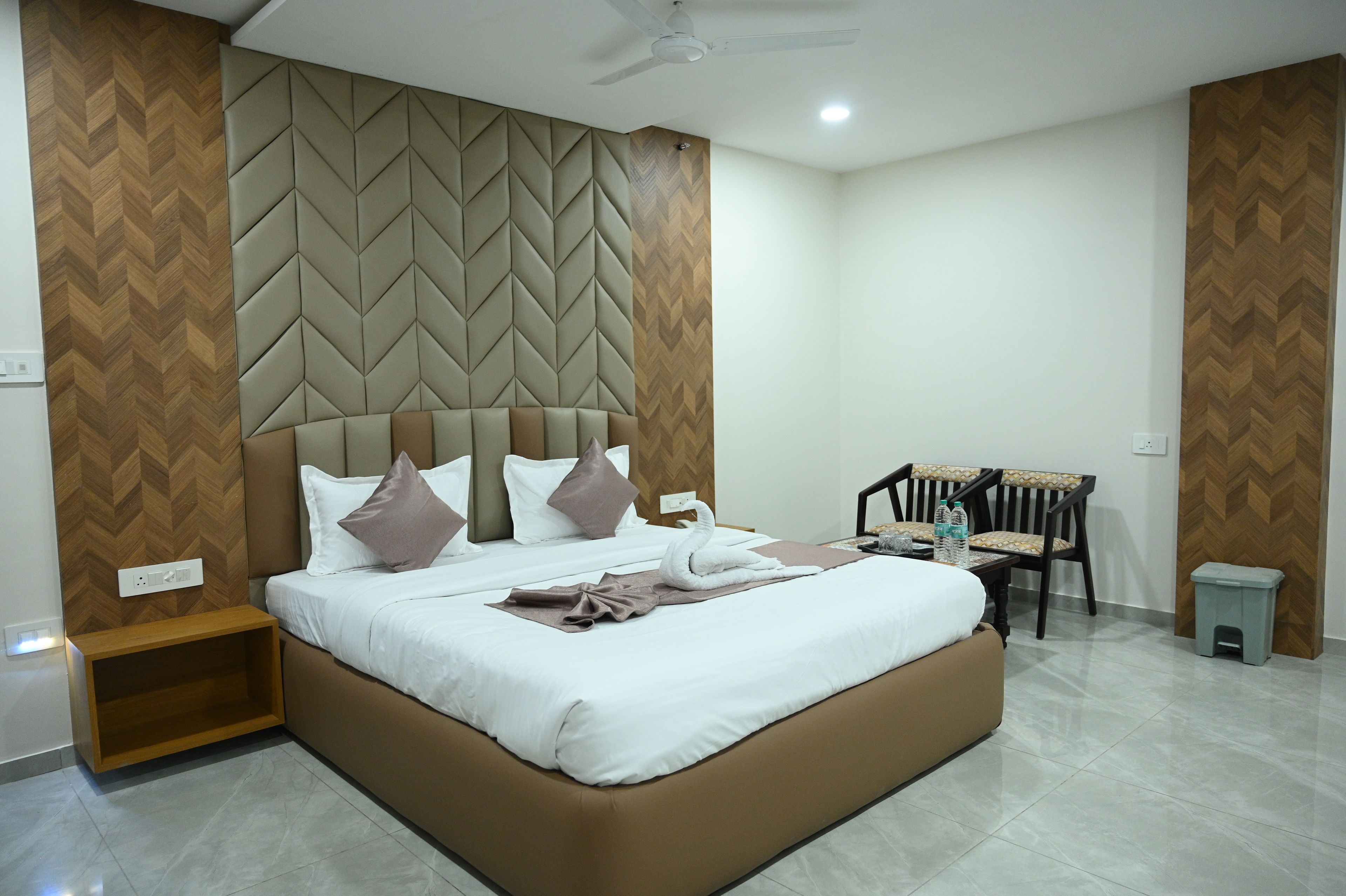 A hotel room with a large bed, a wooden headboard, a nightstand, a chair, and a small stool.
