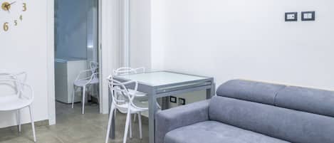 A modern living room with a grey sofa, a dining table with chairs, and a wall clock.