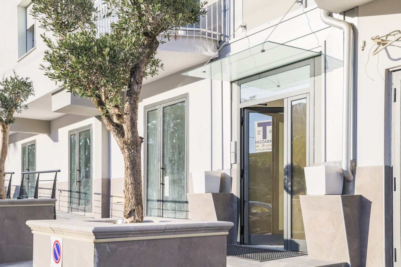 L’entrée d’un hôtel, avec une porte en verre, un arbre au premier plan et un panneau indiquant qu’il est interdit de se garer.