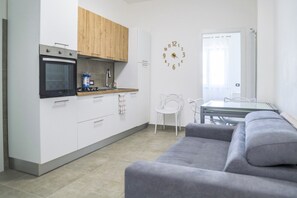 A modern kitchen with white cabinets, a wooden upper section, and a built-in oven. There's a dining area with a glass table and white chairs, and a grey sofa in the foreground. - Royal Gardens - San Leucio (Caserta)