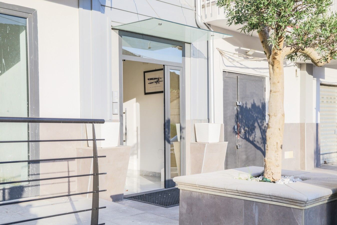 Une entrée moderne avec une porte en verre, une rambarde métallique et un arbre planté dans un bac.