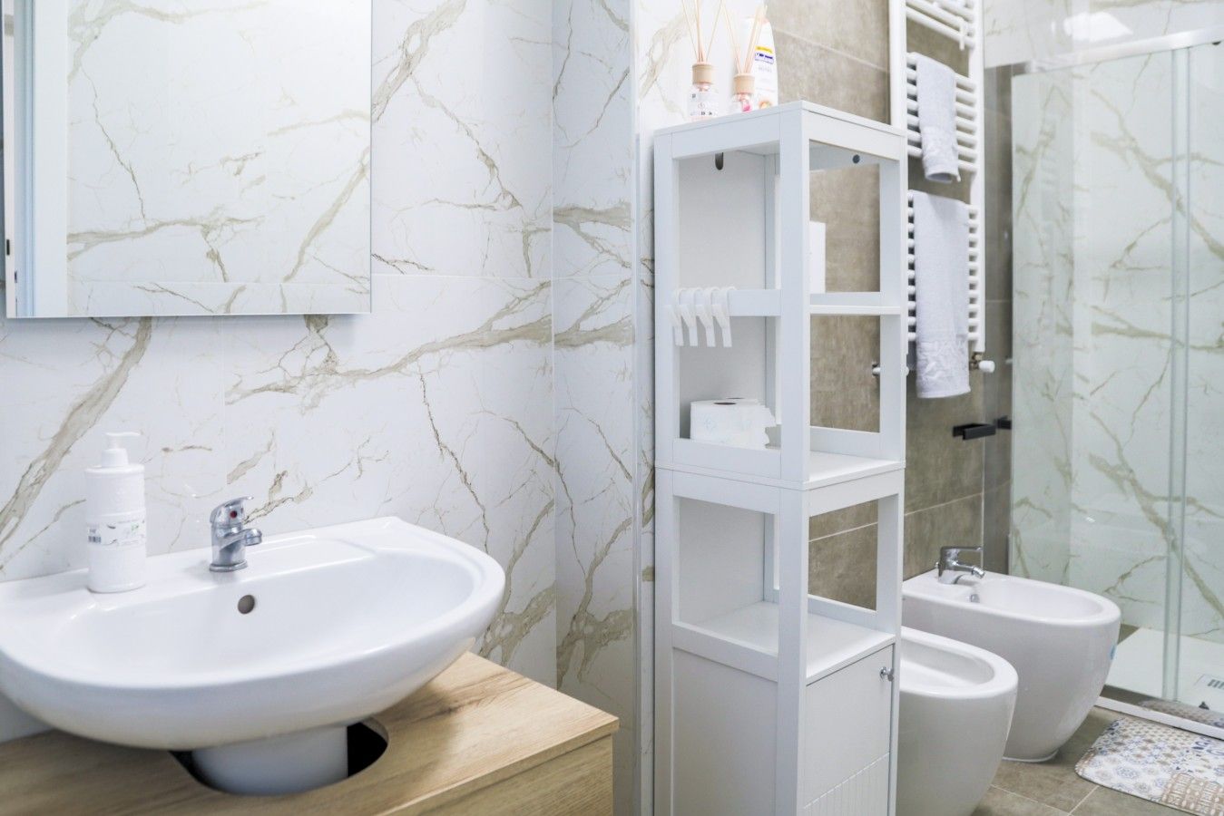 A modern bathroom with a marble wall, a white sink, a toilet, a bidet, and a glass shower enclosure.