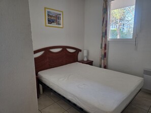 A bedroom with a bed, a nightstand, a lamp, a framed picture on the wall, and a window with a view of buildings and trees. - Air-conditioned apartment with pool near the coves (Fréjus)