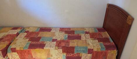 Une petite chambre avec un lit simple, une tête de lit en bois, une courtepointe à motifs et un revêtement de sol carrelé.