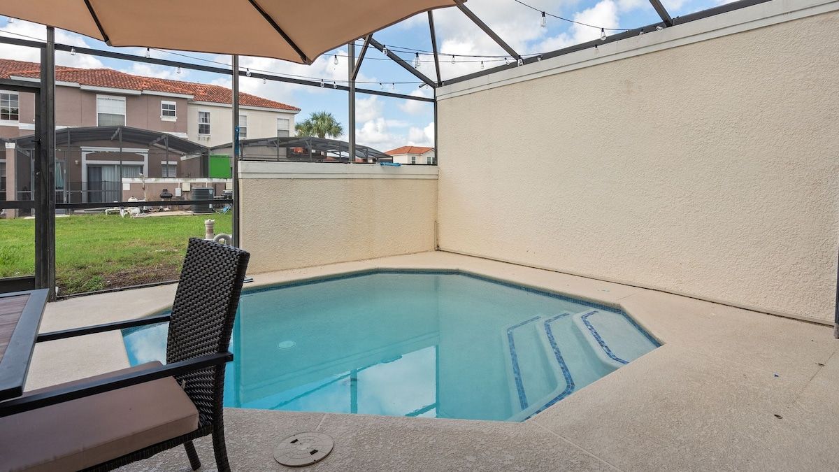 Une piscine entourée de parois en verre, une table avec une chaise, et une vue sur les maisons voisines.